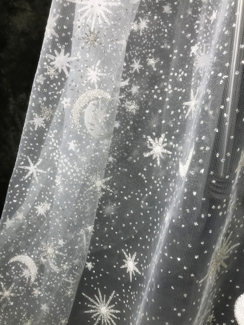 May include: White sheer fabric with a celestial design. The fabric features embroidered stars, moons, and scattered silver glitter, creating a shimmering, starry night effect. Ideal for crafting or decorative purposes.