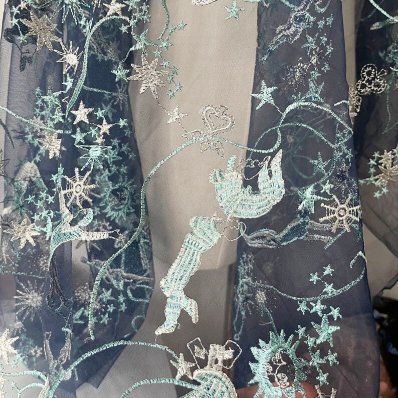 May include: Close-up of a sheer black fabric with a delicate embroidered design of stars, vines, and abstract shapes in silver and light blue.