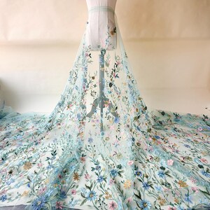May include: A light blue tulle fabric with embroidered floral designs in shades of blue, pink, and yellow. The fabric is draped over a mannequin, showcasing the intricate floral patterns. This fabric is suitable for bridal wear and other special occasion garments.