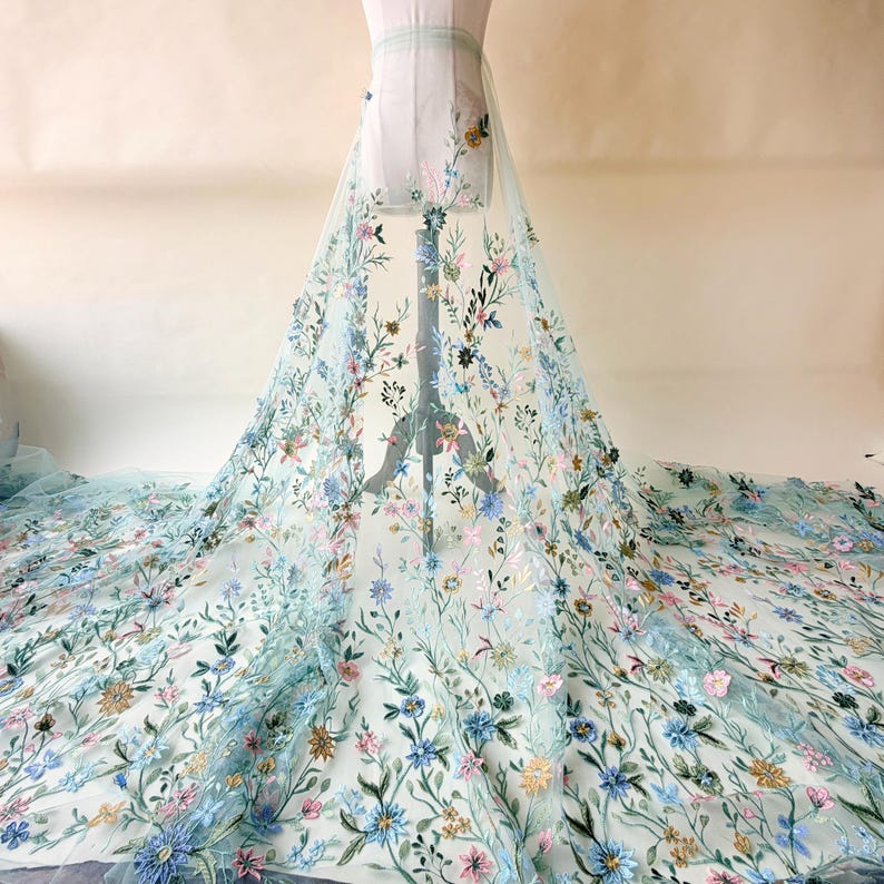 May include: A light blue tulle fabric with embroidered floral designs in shades of blue, pink, and yellow. The fabric is draped over a mannequin, showcasing the intricate floral patterns. This fabric is suitable for bridal wear and other special occasion garments.