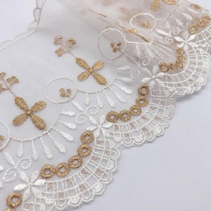 May include: Close-up of white lace trim with gold embroidered floral and cross designs. The lace has a scalloped edge and is likely used for sewing or crafting projects. The delicate details and colors suggest a vintage or elegant aesthetic.