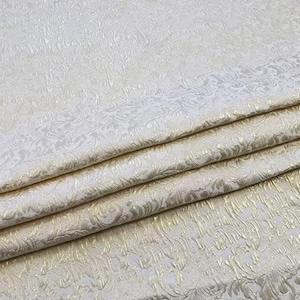 May include: Close-up of folded fabric with a detailed, embossed floral pattern. The fabric is a creamy white color with subtle gold accents, creating a luxurious and textured appearance. The folds reveal layers of the material.