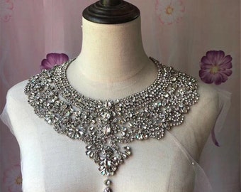 Puede incluir: Un collar de gargantilla grande de color plateado con pedrería y un colgante en forma de lágrima. El collar está hecho de muchas pequeñas piedras de cristal transparentes que están dispuestas en un patrón geométrico.