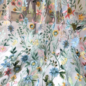 May include: Sheer fabric with colorful floral embroidery. The design features embroidered flowers in shades of pink, blue, yellow, and green, with delicate stems and leaves. The fabric is semi-transparent, allowing light to pass through.