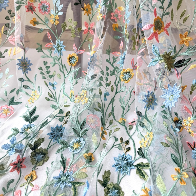 May include: Sheer fabric with colorful floral embroidery. The design features embroidered flowers in shades of pink, blue, yellow, and green, with delicate stems and leaves. The fabric is semi-transparent, allowing light to pass through.