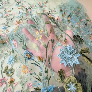 May include: Delicate floral embroidered fabric in shades of blue, pink, yellow, and green. The embroidery features detailed flowers and leaves on a sheer, light green mesh background. The fabric is held up, showcasing the intricate design.