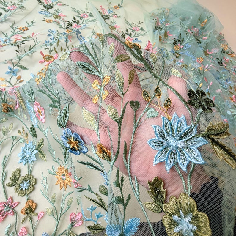 May include: Delicate floral embroidered fabric in shades of blue, pink, yellow, and green. The embroidery features detailed flowers and leaves on a sheer, light green mesh background. The fabric is held up, showcasing the intricate design.
