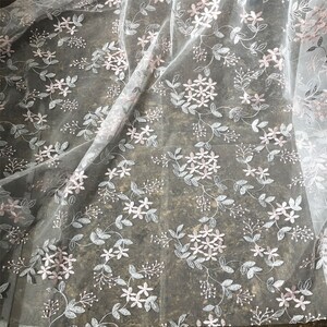 May include: White tulle fabric with a delicate floral pattern in pink and silver. The fabric is sheer and has a soft, flowing texture.