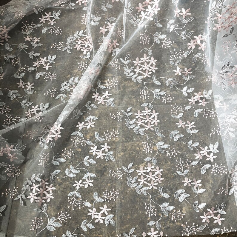 May include: White tulle fabric with a delicate floral pattern in pink and silver. The fabric is sheer and has a soft, flowing texture.