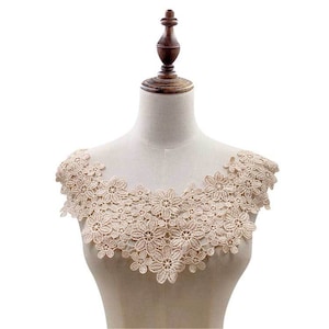 Peut inclure: Un col en dentelle beige avec un motif floral délicat. Le col est présenté sur un mannequin blanc.