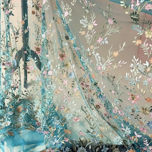 May include: Sheer turquoise fabric with embroidered floral patterns in pink, yellow, and white. The delicate embroidery creates a textured effect, with the flowers and leaves scattered across the fabric. The fabric is lightweight and flowing, suitable for apparel or home decor.