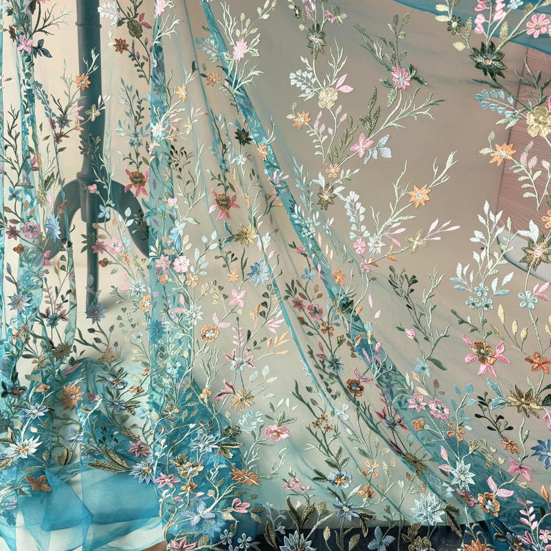May include: Sheer turquoise fabric with embroidered floral patterns in pink, yellow, and white. The delicate embroidery creates a textured effect, with the flowers and leaves scattered across the fabric. The fabric is lightweight and flowing, suitable for apparel or home decor.