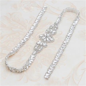 May include: A silver bridal sash with a delicate crystal design. The sash features a central cluster of crystals and two smaller clusters on either side. The sash is made of a thin, flexible material and is perfect for adding a touch of sparkle to any wedding dress.
