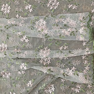 May include: White sheer fabric with a delicate floral embroidery pattern. The flowers are pink and the leaves are green.