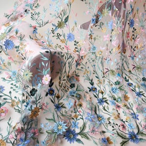 May include: Delicate floral embroidered fabric featuring a variety of colorful flowers and green leaves. The flowers are in shades of blue, pink, yellow, and brown, creating a vibrant and detailed pattern. The fabric has a sheer, light pink background.