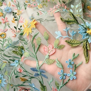 May include: Close-up of embroidered floral fabric. The sheer material features colorful flowers in shades of yellow, blue, pink, and green. The delicate embroidery creates a textured, three-dimensional effect, perfect for apparel or craft projects.