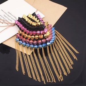 May include: A decorative shoulder piece with a spiky design and gold chain tassels. The piece features a gradient of colours, including black, pink, gold, and blue. It is placed on a brown envelope.
