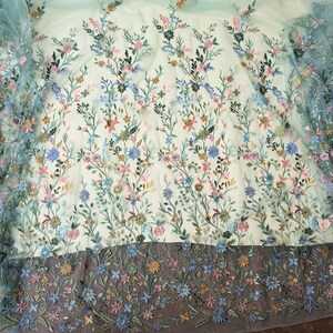 May include: Delicate floral embroidered fabric in shades of blue, pink, and gold on a light background. The design features detailed flowers and leaves, creating a textured, three-dimensional effect. The bottom edge has a darker border with more floral embroidery.