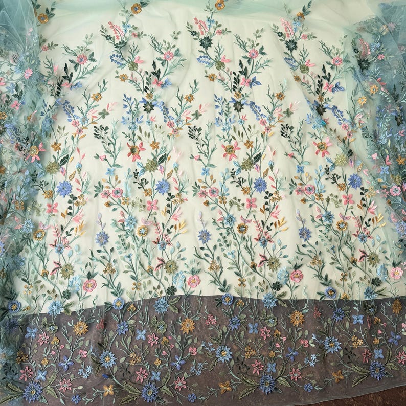 May include: Delicate floral embroidered fabric in shades of blue, pink, and gold on a light background. The design features detailed flowers and leaves, creating a textured, three-dimensional effect. The bottom edge has a darker border with more floral embroidery.
