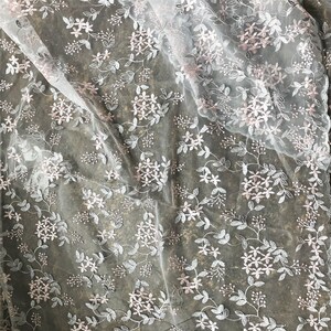 May include: White sheer fabric with a delicate floral embroidery pattern in pink and silver. The fabric is draped and shows the texture of the embroidery.