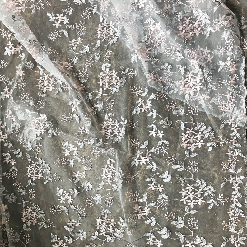 May include: White sheer fabric with a delicate floral embroidery pattern in pink and silver. The fabric is draped and shows the texture of the embroidery.