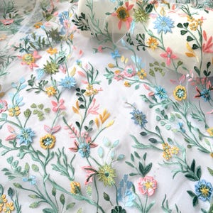 May include: Delicate floral embroidered fabric featuring a repeating pattern of colorful flowers and leaves. The embroidery includes pink, yellow, blue, and green blossoms with detailed stitching. The fabric is a sheer white material.