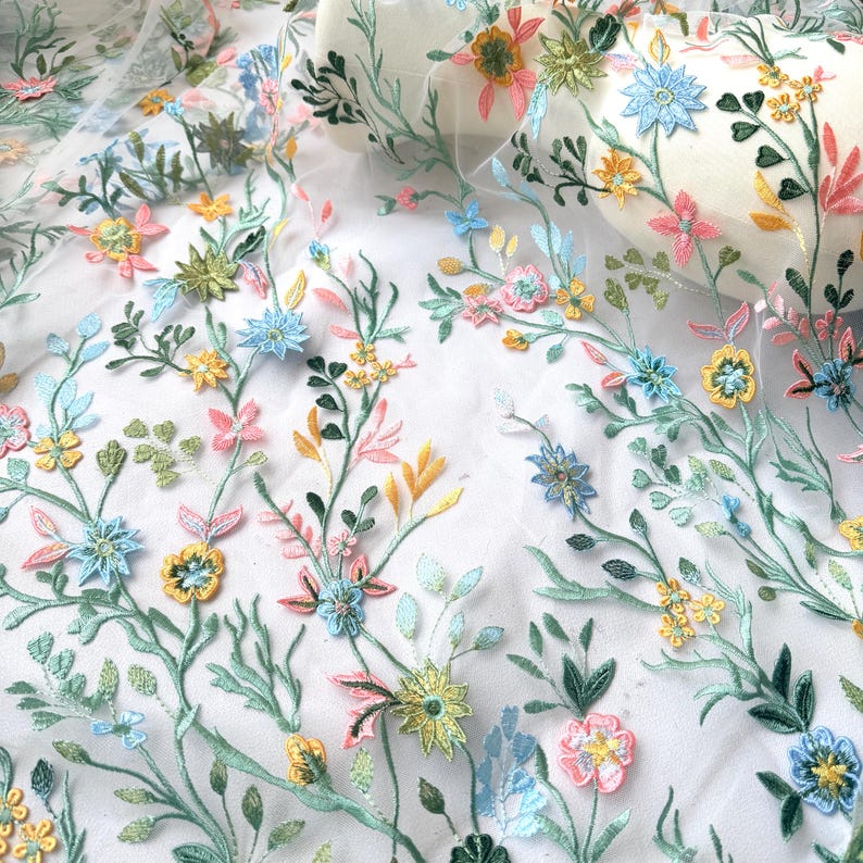 May include: Delicate floral embroidered fabric featuring a repeating pattern of colorful flowers and leaves. The embroidery includes pink, yellow, blue, and green blossoms with detailed stitching. The fabric is a sheer white material.