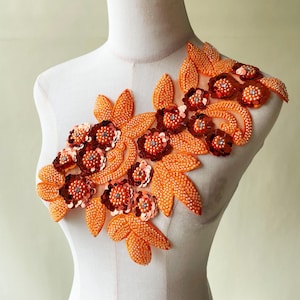 Op de afbeelding: Een oranje bloemenapplicatie met kralen en pailletten. Het ontwerp heeft meerdere bloemen met donkerrode centra en zilveren accenten, omgeven door oranje gebladerte en stelen met kralen. De applicatie wordt op een mannequin getoond.