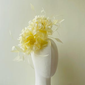 Peut inclure: Coiffe florale jaune pâle avec des plumes délicates. La coiffe présente deux grandes fleurs superposées et plusieurs petites plumes vaporeuses. Les fleurs sont d'un jaune doux et les plumes sont d'une nuance de jaune plus claire.