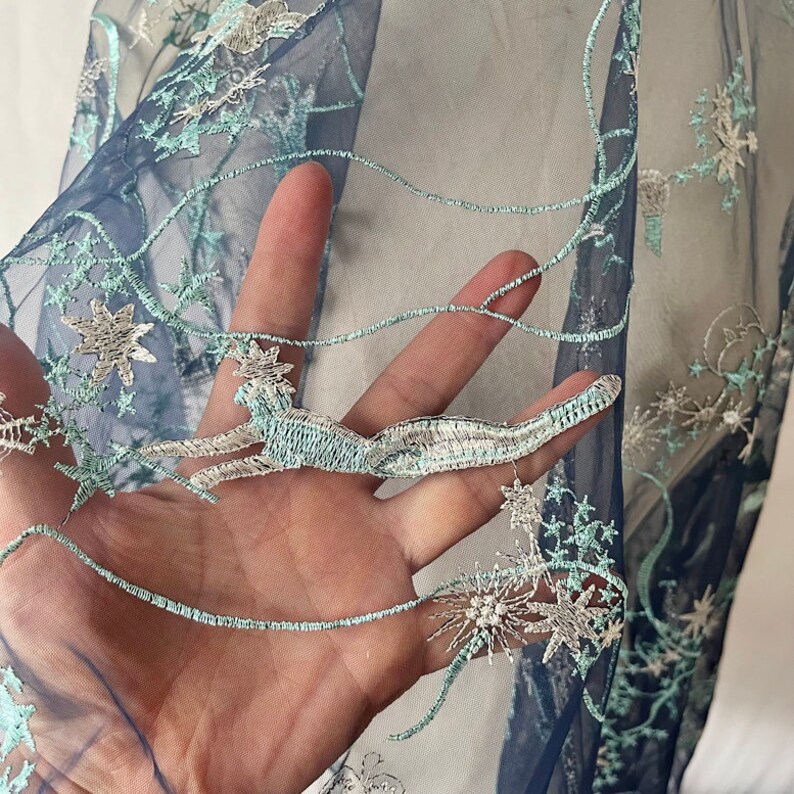 May include: Close-up of a light blue sheer fabric with silver and light blue embroidered stars and floral designs.