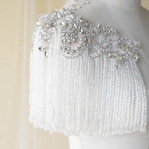Peut inclure: Une épaule de mariée en perles blanches avec un ornement en cristal et strass. La pièce a un design en frange et est parfaite pour ajouter une touche d'éclat à une robe de mariée.