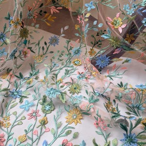 May include: Delicate floral embroidered fabric featuring a variety of colorful flowers and leaves. The embroidery includes blue, yellow, pink, and green hues, creating a vibrant and detailed design. The fabric appears to be sheer, allowing light to pass through.