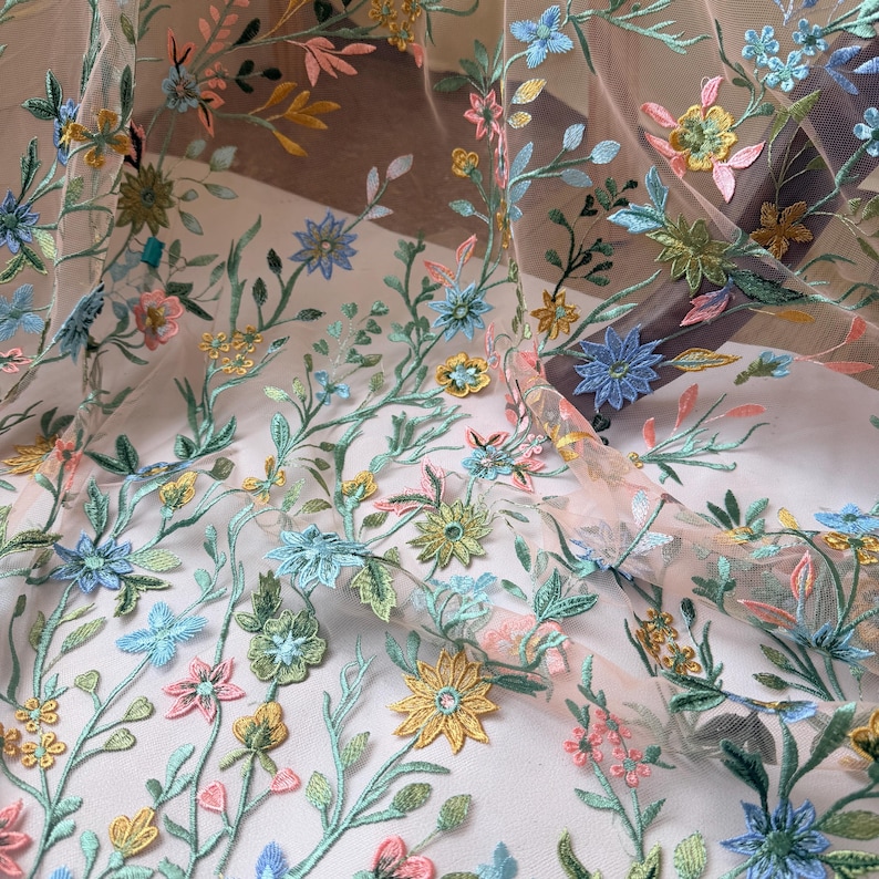 May include: Delicate floral embroidered fabric featuring a variety of colorful flowers and leaves. The embroidery includes blue, yellow, pink, and green hues, creating a vibrant and detailed design. The fabric appears to be sheer, allowing light to pass through.