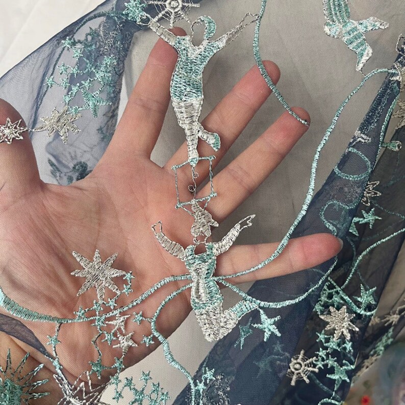 May include: Blue and silver embroidered tulle fabric with a design of stars and figures on a trapeze. The fabric is sheer and delicate.