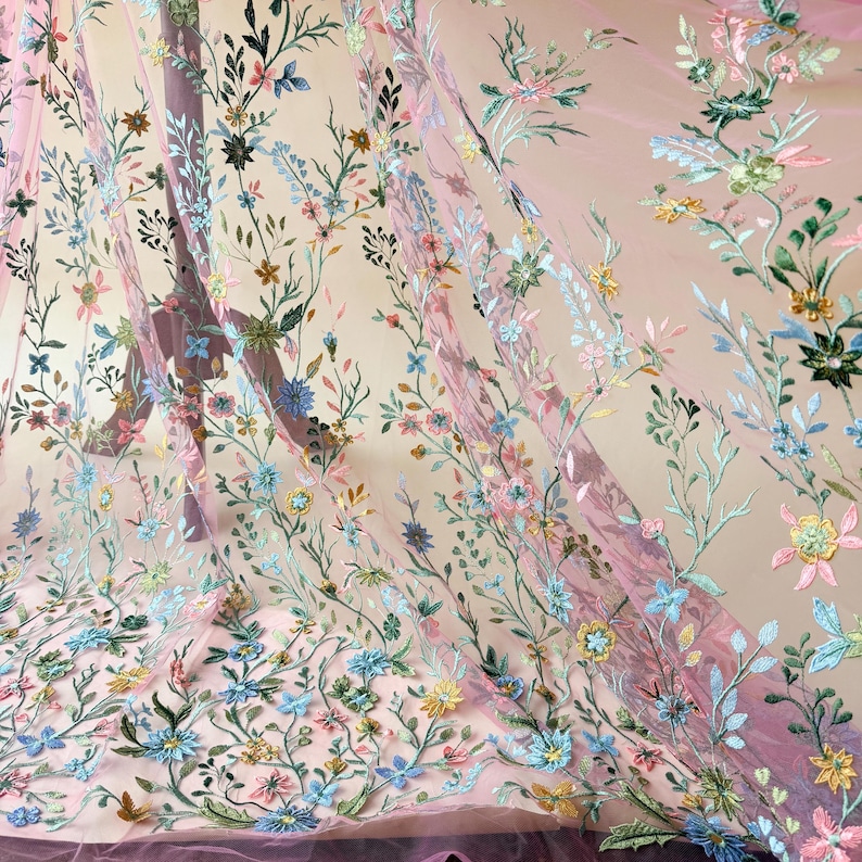 May include: Sheer pink fabric with embroidered floral patterns. The embroidery features a variety of colorful flowers in shades of blue, yellow, green, and pink, with delicate green stems and leaves. The fabric is lightweight and flowing, ideal for apparel or decorative projects.