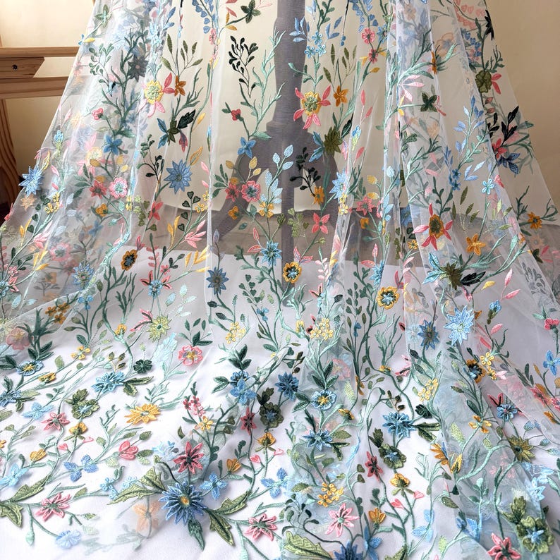 May include: Sheer white fabric with colorful floral embroidery. The embroidery features a variety of flowers in shades of blue, pink, yellow, and green, with delicate green leaves and stems. The fabric is draped, showcasing the intricate details.