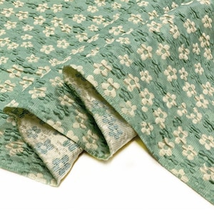 May include: A close-up of a green fabric with a white floral pattern. The fabric is textured and has a slightly raised design.