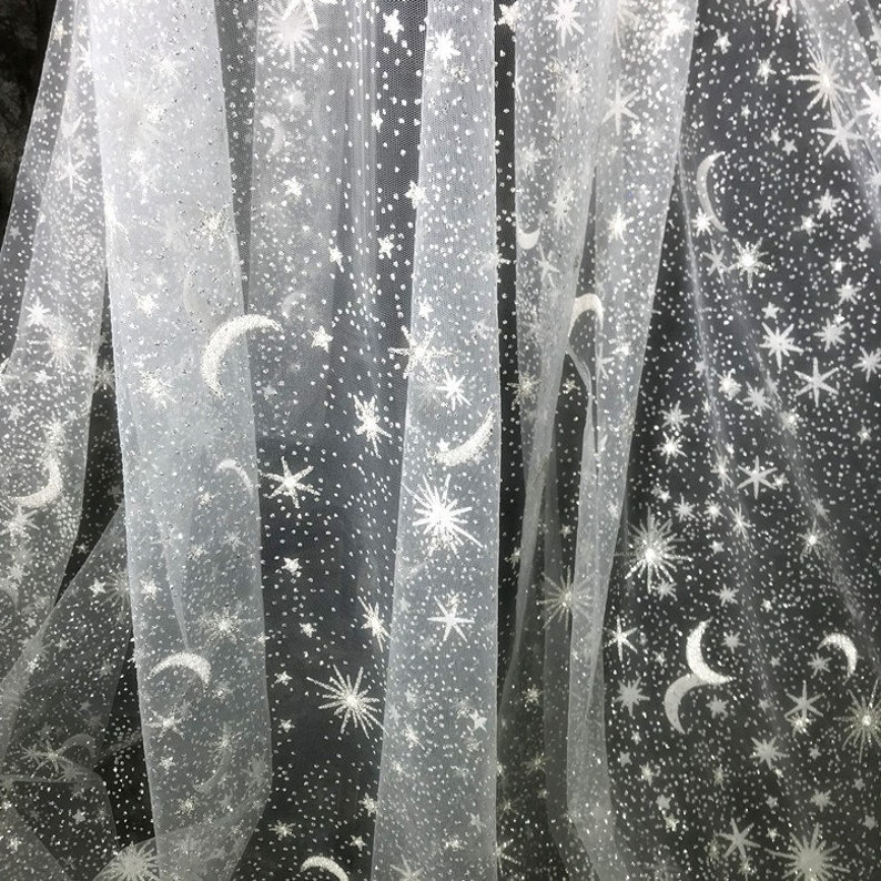 May include: White sheer fabric with a celestial pattern of silver stars, moons, and dots. The fabric is draped, creating soft folds and shadows. The design is evenly distributed across the material, suitable for apparel or decorative projects.