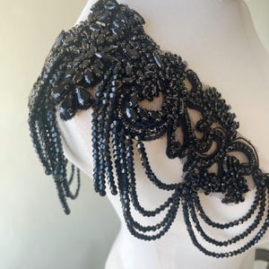 Könnte beinhalten: Schwarze Perlen-Schulterverzierung mit Fransen-Design. Die Verzierung hat ein florales Muster und besteht aus schwarzen Perlen und Pailletten.