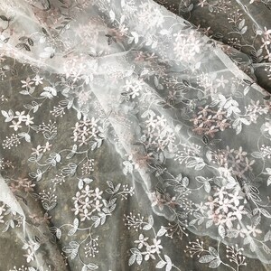 May include: White tulle fabric with delicate floral embroidery in a light pink color. The fabric has a soft, airy feel and is perfect for creating romantic and elegant garments.