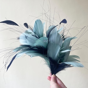 May include: A cluster of decorative feathers in shades of teal and navy blue. The feathers are arranged in a bouquet-like shape, with long, thin, dark strands extending outwards. The feathers are held together at the base.