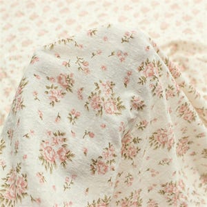 May include: Close-up of a white fabric with a delicate floral pattern. The design features clusters of pink roses and smaller pink buds with green leaves. The fabric is soft and slightly textured, with a vintage aesthetic.