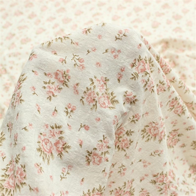 May include: Close-up of a white fabric with a delicate floral pattern. The design features clusters of pink roses and smaller pink buds with green leaves. The fabric is soft and slightly textured, with a vintage aesthetic.