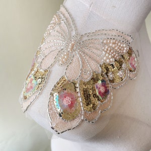 Può includere: Un'applicazione delicata a forma di farfalla con paillettes dorate, perline iridescenti e dettagli di perle. L'applicazione è realizzata in un tessuto trasparente ed è perfetta per aggiungere un tocco di brillantezza a vestiti o accessori.