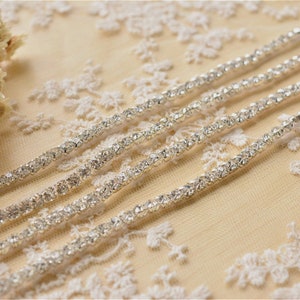 Puede incluir: Primer plano de una cinta de adorno de strass plateado, con una fila de pequeños strass brillantes. La cinta está colocada sobre un fondo de tela de encaje blanco.