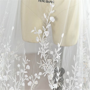May include: White floral lace fabric with delicate embroidery and sequins. The lace features a repeating pattern of small flowers and leaves.