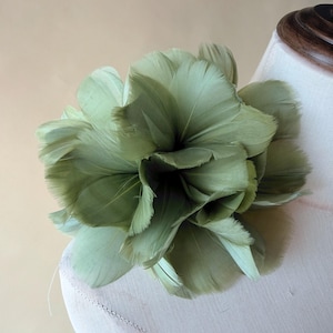 May include: A large, sage green feather flower, possibly a brooch or hair accessory. The flower is made of overlapping feathers, creating a full, textured appearance. The accessory is displayed on a mannequin, showcasing its shape and design.