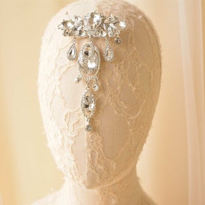 Peut inclure: Un peigne de cheveux de mariée en argent et cristal avec un motif en forme de larme. Le peigne est sur une tête de mannequin en dentelle blanche.