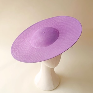Puede incluir: Un sombrero grande de color lavanda con ala ancha y copa redonda. El sombrero se exhibe sobre una cabeza de maniquí blanca sobre un fondo neutro. La textura del sombrero parece ser tejida, lo que sugiere un material ligero.