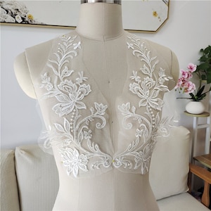 以下が含まれることがあります： 繊細なビーズをあしらった白い花のレースアップリケ。ウェディングドレス、ブライダルアクセサリー、その他のクラフトプロジェクトにエレガントなアクセントを加えるのに最適です。